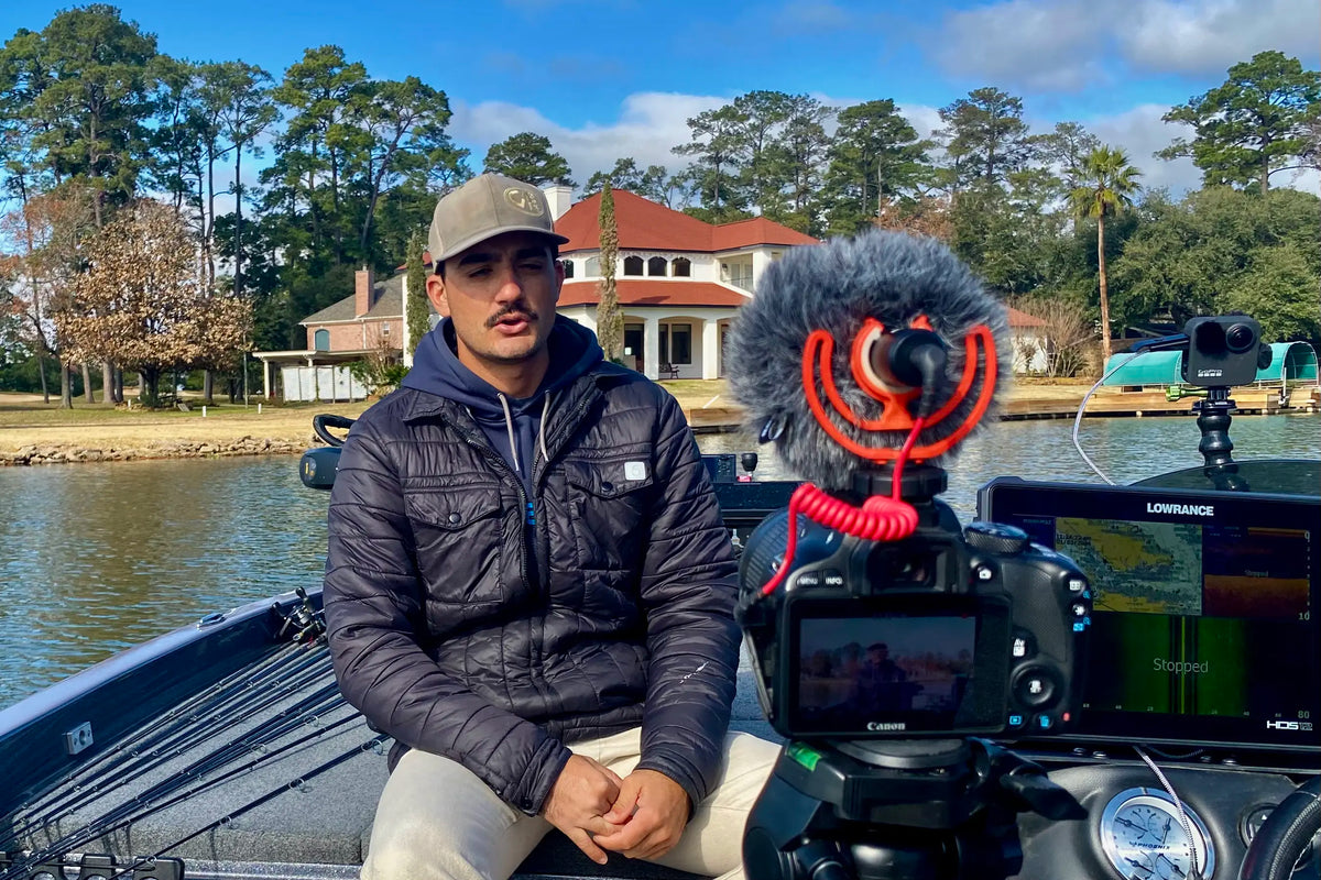 Man on a boat with fishing equipment and camera, surrounded by trees and houses.