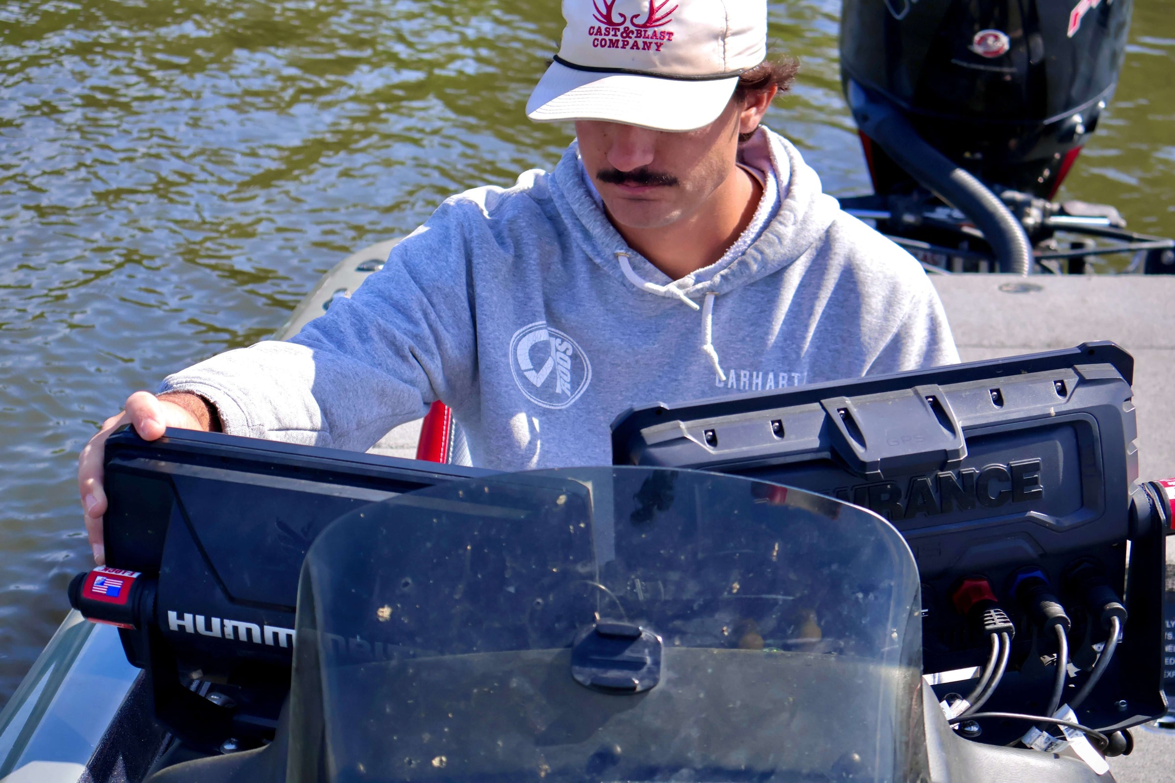 Person operating dual fish finders on a boat in a lake, wearing a gray hoodie and white cap.