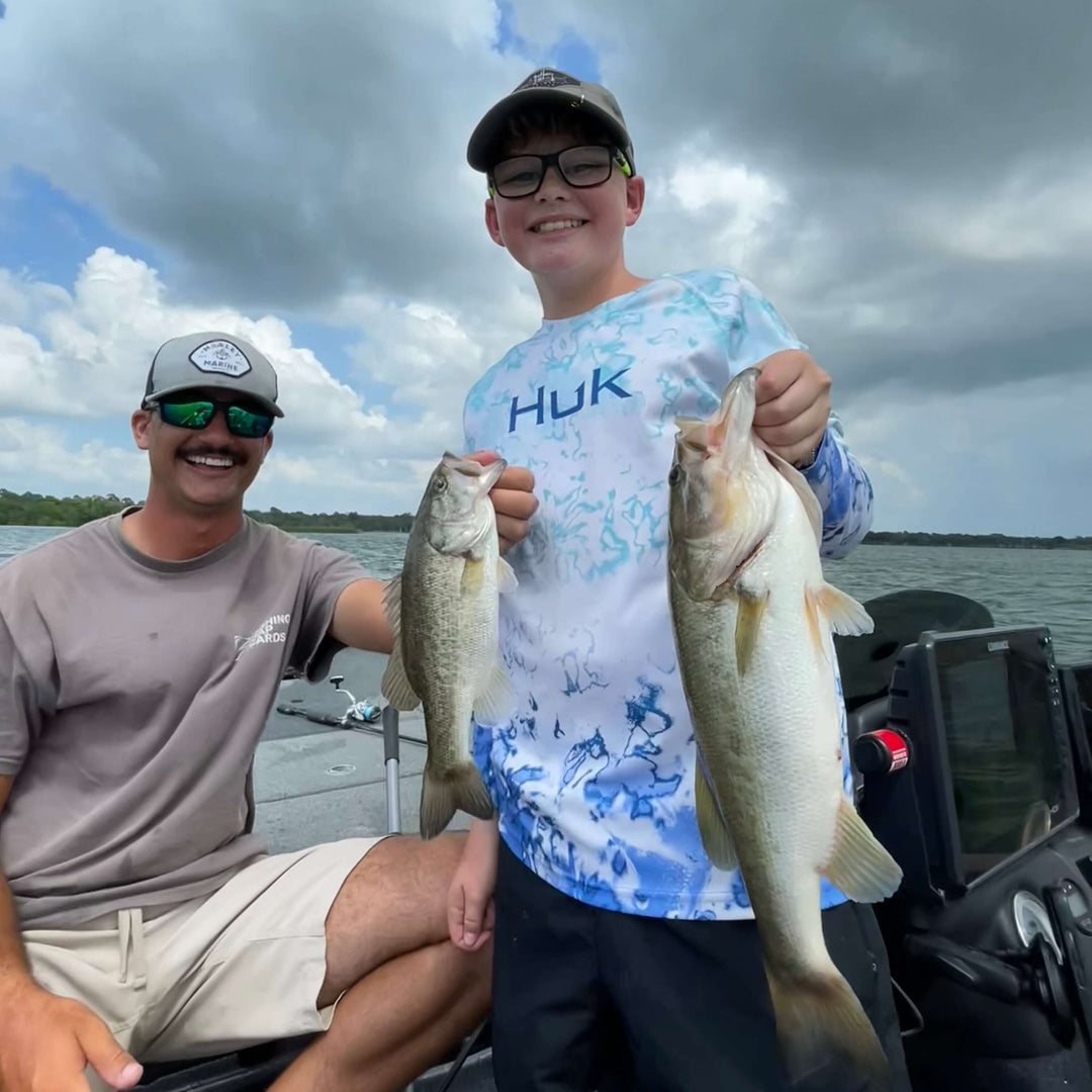 Father and son proudly display their catch of two bass fish on a boat under cloudy skies.