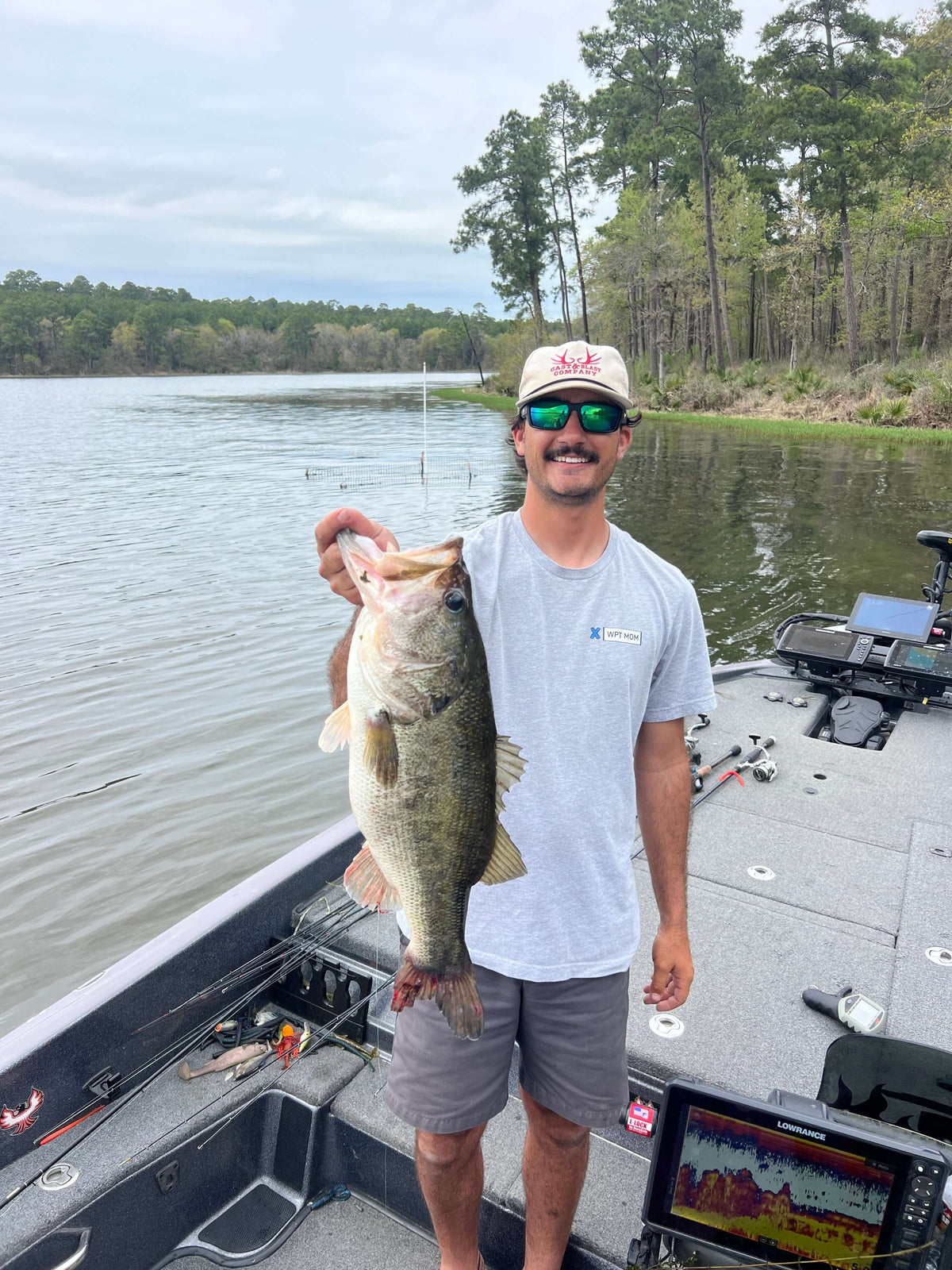 Angler proudly holding a large bass fish on a boat in a serene lake setting, surrounded by lush greenery and calm waters.