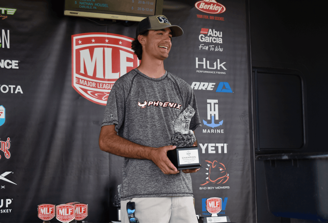 Angler holding a trophy at Major League Fishing event, smiling on stage with sponsor banners in the background.