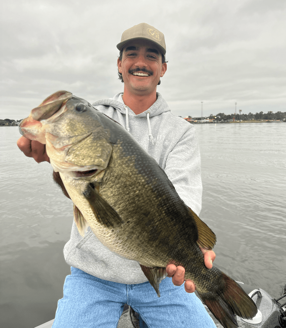 Man proudly holding a large bass fish on a boat in a lake, wearing a cap and hoodie, showcasing a successful fishing trip.