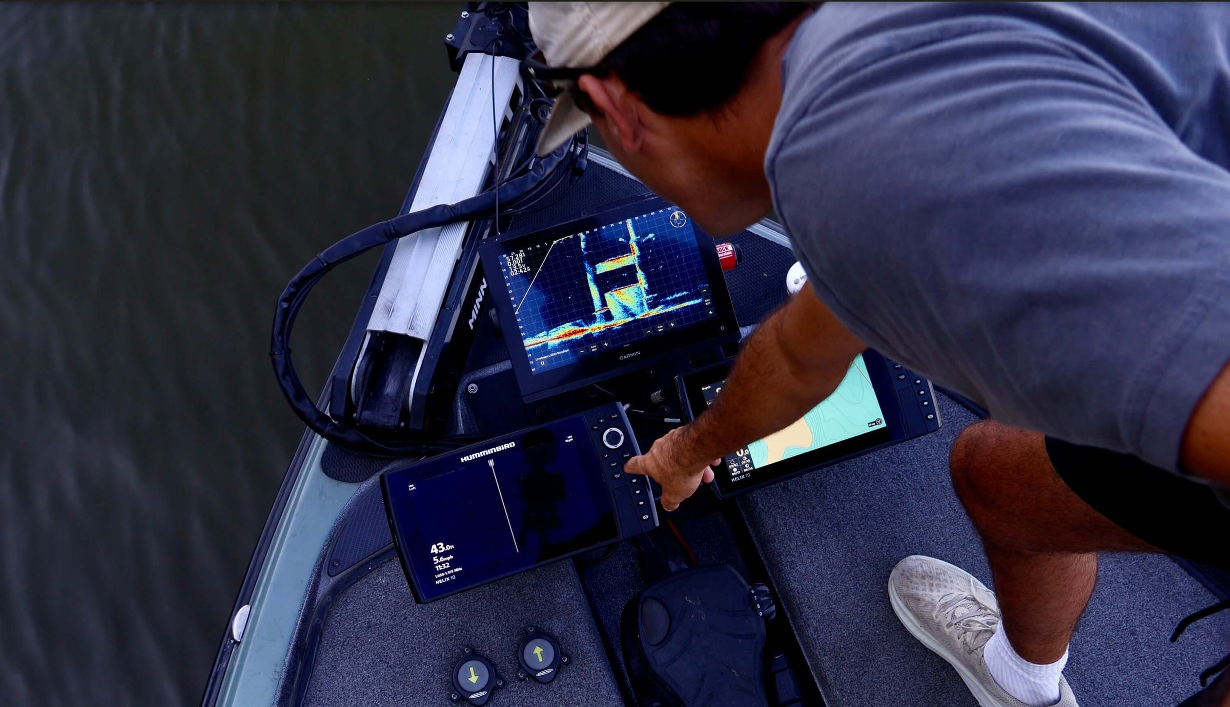 Person using a fish finder on a boat, pointing at multiple screens displaying sonar and fishing data.