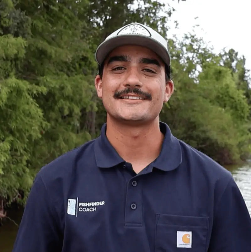 Man wearing a navy blue shirt with a logo, standing outdoors with greenery in the background